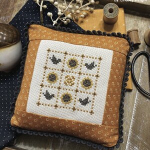 May include: A white pillow with a cross-stitch design of a nine-patch pattern featuring sunflowers and birds. The pillow is surrounded by a brown fabric with a white polka dot pattern and a black and white trim. The text "SEPTEMBER NINE PATCH" is at the top of the image and "PRIMROSE COTTAGE STITCHES" is at the bottom.