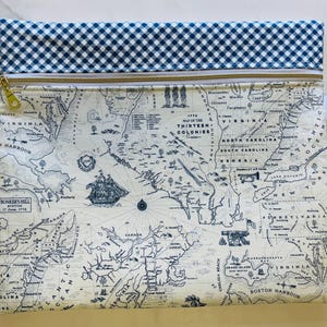 May include: A rectangular pouch featuring a map of the thirteen colonies in blue and white. The top edge has a blue and white gingham pattern. A gold zipper and a small tag are visible. The map includes illustrations of a ship and text that reads "Bunker's Hill Boston".