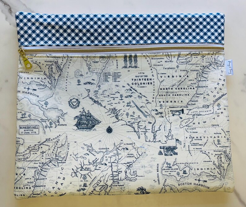 May include: A rectangular pouch featuring a map of the thirteen colonies in blue and white. The top edge has a blue and white gingham pattern. A gold zipper and a small tag are visible. The map includes illustrations of a ship and text that reads "Bunker's Hill Boston".