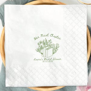 May include: White square paper napkin with a diamond pattern, featuring the text "Her Next Chapter" and a floral and book design in green. The napkin is on a wooden plate, with a gold fork and knife.