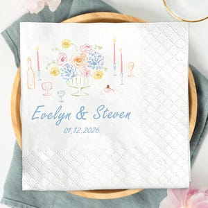 May include: A white napkin with a floral illustration and the names "Evelyn & Steven" with the date 01.12.2026 in blue. The napkin is on a wooden plate with a gold fork and knife. A glass and pink flowers are also in the image.
