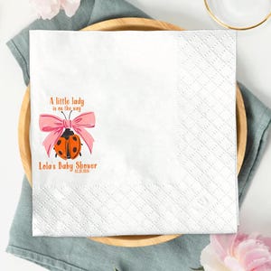 May include: A white paper napkin with a pink bow and a ladybug illustration, with the text "A little lady is on the way" and "Lola's Baby Shower". The napkin is on a wooden plate with a gold fork and knife.