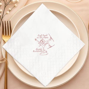 May include: White square paper napkin with a diamond pattern, featuring a pink illustration of a skier in a martini glass. Text reads "Last toast on the slopes" and "Lucy's Bachelorette". The napkin is placed on a white plate with gold cutlery.