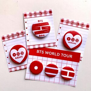 May include: A collection of red and white BTS World Tour pinback buttons. The buttons feature the BTS logo, the word "ARMY", and other designs. The buttons are displayed on small cards with a red and white checkered border.