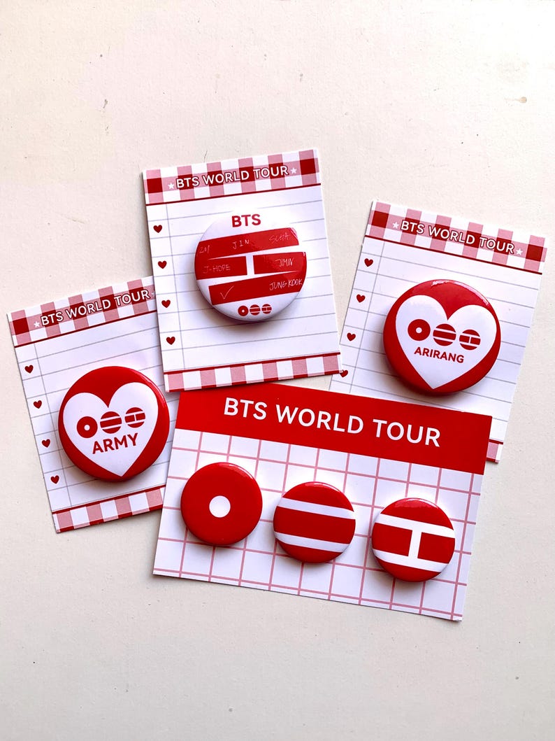May include: A collection of red and white BTS World Tour pinback buttons. The buttons feature the BTS logo, the word "ARMY", and other designs. The buttons are displayed on small cards with a red and white checkered border.