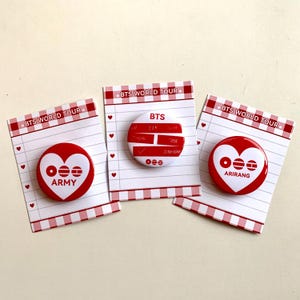 May include: Three red and white pinback buttons with the text "BTS WORLD TOUR" on the top. Two buttons have a heart design with the text "ARMY" and "ARIRANG". The third button has the text "BTS" and other names.