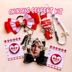 Op de afbeelding: Een verzameling rode en witte accessoires, waaronder een haarstrik, een lanyard, een kralenketting en een fotolijst. De items bevatten de tekst "ARIRANG CONCERT KIT" en "BTS WORLD TOUR". Het kleurenschema is voornamelijk rood en wit.
