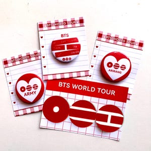 May include: A collection of red and white BTS World Tour buttons and cards. The buttons feature the BTS logo, names, and heart designs. The cards have a grid pattern and the text "BTS WORLD TOUR".