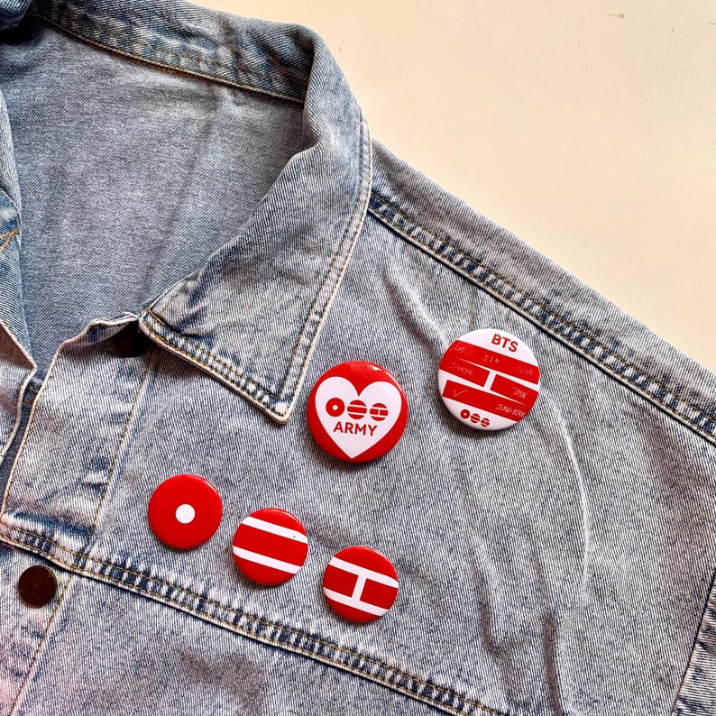 May include: A collection of red and white pinback buttons with various designs, including a heart with "ARMY" text and a button with "BTS" text. The buttons are arranged on a light blue denim jacket.
