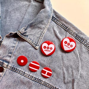 May include: A collection of red and white button pins with various designs, including a heart shape with the text "ARMY" and "ARIRANG". The pins are displayed on a light blue denim jacket, showcasing a casual style.