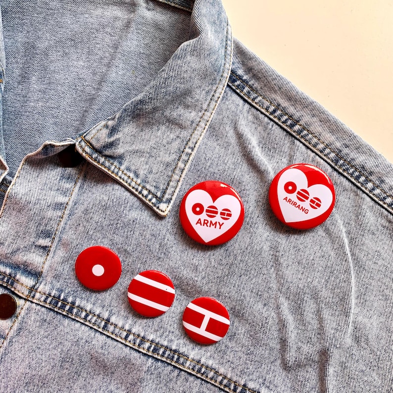 May include: A collection of red and white button pins with various designs, including a heart shape with the text "ARMY" and "ARIRANG". The pins are displayed on a light blue denim jacket, showcasing a casual style.