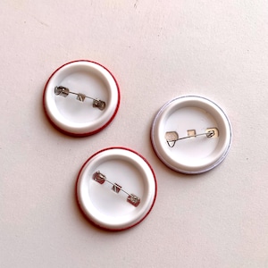 May include: Three round pinback buttons with safety pin closures. Two buttons have a red border and one has a silver border. The interior of each button is white.