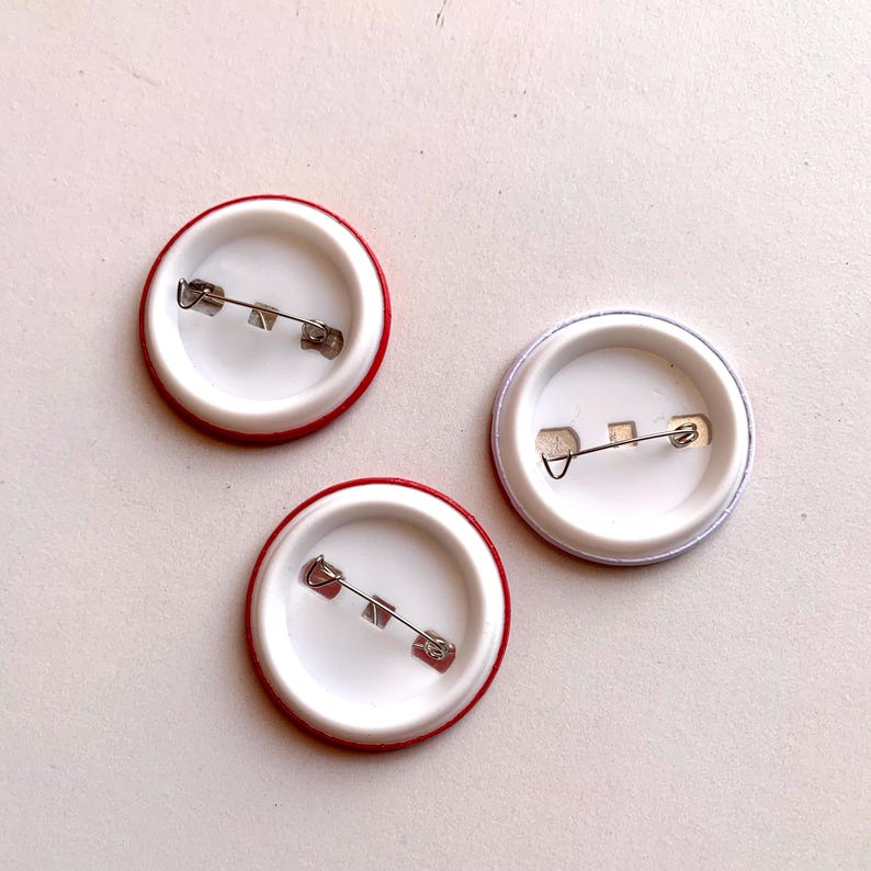 May include: Three round pinback buttons with safety pin closures. Two buttons have a red border and one has a silver border. The interior of each button is white.