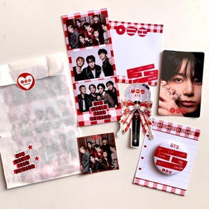 Peut inclure: Divers produits dérivés BTS, comprenant des cartes photo, un bâton lumineux avec un nœud rouge et blanc, des autocollants et un bloc-notes. Les articles présentent le logo BTS et des images des membres du groupe. Fond blanc.