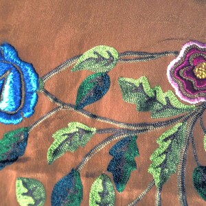 May include: Close-up of embroidered floral design featuring a blue rose and a purple flower with yellow center. Green and blue leaves are also embroidered on a brown fabric background. The embroidery is detailed and textured.