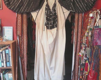 May include: A cream-colored, long-sleeved dress with black and white embroidered details. The sleeves are wide and patterned with diagonal stripes. The neckline and hem are also decorated with black embroidery. The dress is hanging in a doorway.