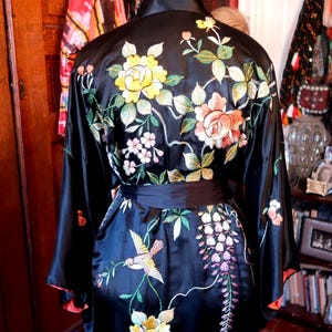 May include: Black silk kimono with floral embroidery in yellow, pink, and white. The kimono features a bird and wisteria design on the front. The sleeves have a red trim. A black belt cinches the waist.