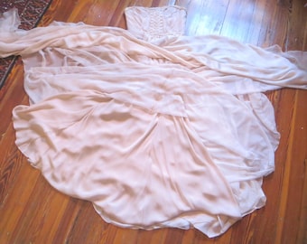 May include: A pale pink strapless gown featuring a fitted bodice with vertical lace-up detailing and a voluminous, flowing skirt made of layered, sheer fabric that spreads out across the floor in a wide, circular arrangement of soft folds.