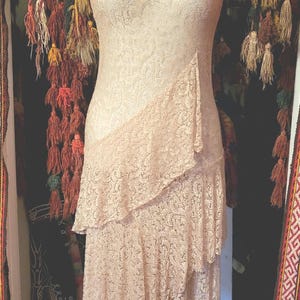 May include: A sleeveless, knee-length dress in a pale peach lace fabric. The garment features a round neckline and a layered, asymmetrical skirt design with cascading ruffles that start from the left hip and extend down to the hemline.