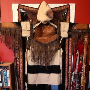 May include: A handcrafted, fringed leather and wool garment with a hooded design. The garment features a brown leather chest piece, cream-colored wool sleeves, and a cream and black striped lower section. The hood is cream-colored with decorative stitching.