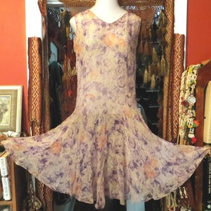 May include: A sleeveless, vintage-style dress with a V-neckline. The dress features a flowing, flared skirt and is adorned with a floral pattern in shades of purple, peach, and cream. The fabric appears sheer and lightweight.