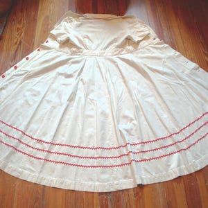 May include: Cream-colored vintage dress with a button-down front and a full skirt. The dress features red embroidered trim along the hem and red buttons. The dress is laid out on a wooden floor.