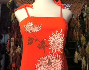 May include: A sleeveless, orange dress with white chrysanthemum floral print. The dress has tie straps and a straight neckline. The floral design includes brown stems and leaves. The dress is displayed on a mannequin.