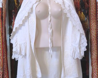 May include: An off-white cape featuring intricate tonal embroidery and eyelet lace trim along the edges. The garment includes a delicate tie closure at the neckline and wide, ruffled sleeves with detailed cutwork patterns throughout the fabric.