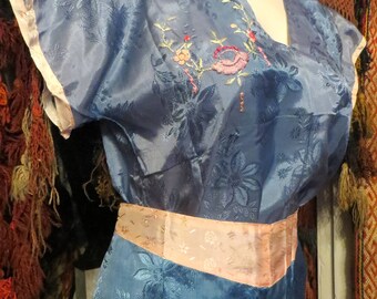 May include: A vintage blue dress with floral embroidery and a peach-colored sash. The dress features short sleeves with peach trim and a V-neckline. The fabric has a subtle floral pattern throughout, giving it a classic, elegant look.