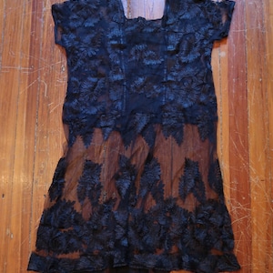May include: A black, sheer, short-sleeved top with intricate floral lace detailing. The lace design is concentrated at the hem and across the bodice, creating a textured, layered effect. The garment is laid flat on a wooden surface.
