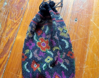 May include: A beaded drawstring bag with a floral pattern in red, orange, purple, and yellow on a black background. The bag has a black drawstring closure and is made of small, colorful beads. The bag is on a wooden surface.