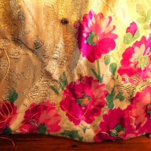 May include: Close-up of a textile with a gold background and vibrant pink floral pattern. The fabric has a metallic sheen and a fringe of gold threads. The flowers have green centers.