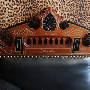 May include: Ornate wooden headboard with black leather upholstery. The headboard features intricate carvings, black accents, and gold-colored stud detailing along the leather edge. The background is a leopard print pattern.