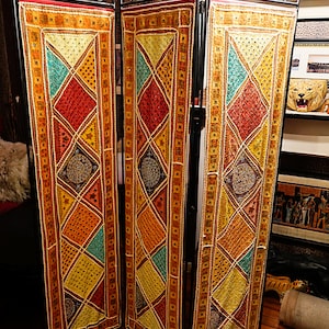 May include: A three-panel room divider with a black metal frame and ornate scrollwork at the top. Each panel features a colorful, geometric design with diamond and square patterns in shades of red, yellow, and teal. The divider stands on a wooden floor.