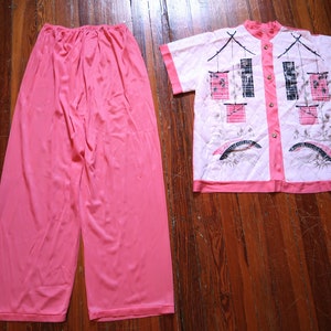 May include: A vintage pink pajama set. The set includes wide-leg pants and a short-sleeved button-up shirt. The shirt features a quilted design with an Asian-inspired illustration in pink, black, and white. The shirt has pink trim and gold buttons.