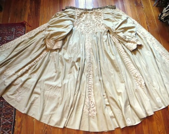 May include: A long, beige-colored garment featuring voluminous sleeves and intricate, cream-colored lace detailing along the bodice, cuffs, and vertical panels. The fabric is gathered at the waist, creating a wide, flowing skirt with lace accents.