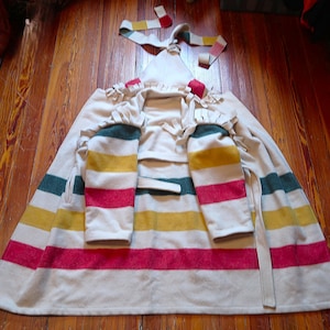 May include: A cream-colored poncho with green, yellow, and red horizontal stripes. The poncho has long sleeves with matching stripes and a fringed collar. A matching tie is at the neck.