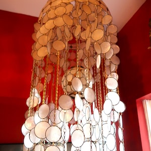 May include: A decorative chandelier with a tiered design, featuring numerous white, oval-shaped elements. The chandelier is suspended from the ceiling and has gold-colored chain accents. The background is a deep red.
