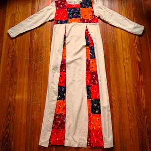 May include: Cream-colored maxi dress with long sleeves and a patchwork bodice and panels. The patchwork features squares of red, orange, navy, and burgundy fabric with floral patterns. The dress is laid out on a wooden surface.