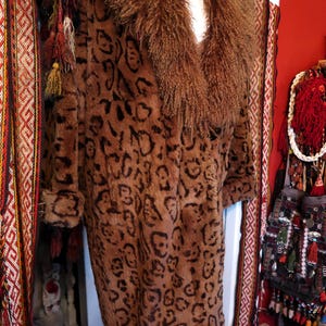 May include: A long, brown leopard print coat with a large, textured collar. The coat is displayed on a mannequin. The background includes decorative textiles and a red wall, creating a vintage aesthetic.