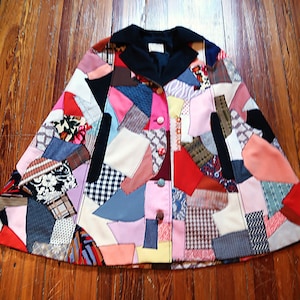 May include: A patchwork cape with a black collar and button closure. The cape is made of various fabric scraps in a variety of colors and patterns, including pink, red, blue, and white. The garment is laid out on a wooden surface.