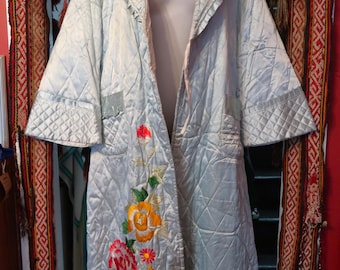 May include: A light blue quilted robe with floral embroidery. The robe features a shawl collar and short sleeves. The embroidery includes red, yellow, and pink flowers. The robe is displayed on a mannequin.