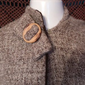 May include: A brown tweed cape with a round wooden clasp. The cape is displayed on a white mannequin head. The fabric has a woven texture. The background features a brown crocheted shawl.