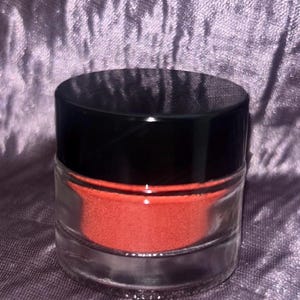 May include: A small, cylindrical clear glass jar filled with a vibrant, matte orange powder. The container is topped with a smooth, solid black circular lid that sits flush against the rim of the glass base, creating a simple, functional design.