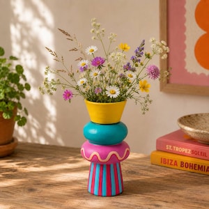 May include: A multi-coloured vase with a yellow top, a teal middle section, and a pink base featuring a wavy line and vertical blue stripes. The vase is filled with a variety of colourful wildflowers including white, pink, yellow, and purple blooms.