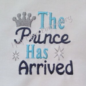 Puede incluir: Diseño bordado sobre un fondo de tela blanco. El diseño presenta una corona plateada, las palabras "The Prince Has Arrived" en letras azules y negras, y dos estrellas plateadas.