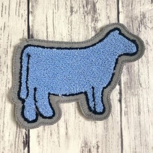 May include: A cow-shaped patch featuring a textured light blue center with a dark navy blue outline, all set against a light gray fabric border. The design is flat and shaped like a bovine silhouette, showcasing a simple and clean aesthetic.