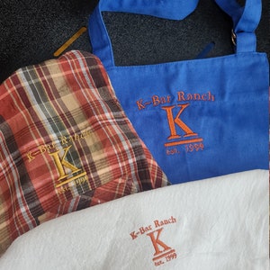 May include: Three different aprons with the same logo embroidered on them. The logo is a stylized "K" with the text "K-Bar Ranch est. 1999" underneath. The aprons are blue, red and white plaid, and white.