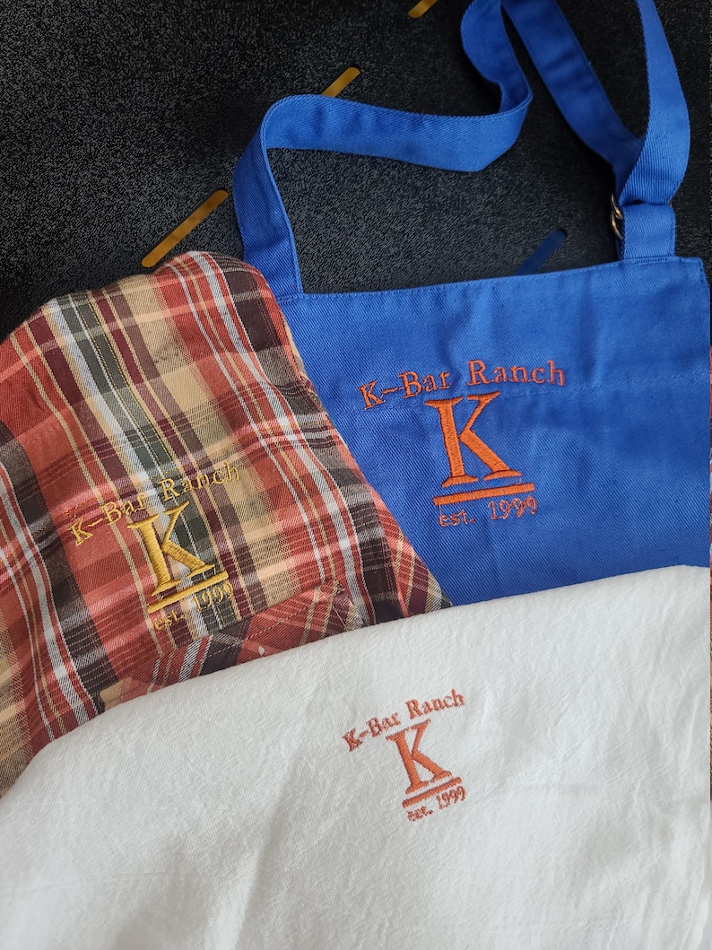 May include: Three different aprons with the same logo embroidered on them. The logo is a stylized "K" with the text "K-Bar Ranch est. 1999" underneath. The aprons are blue, red and white plaid, and white.