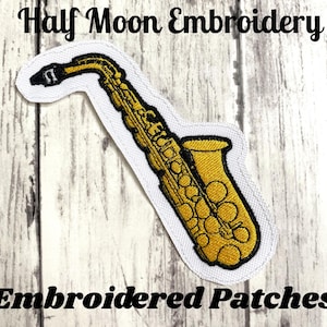 Pode incluir: Patch bordado com um saxofone dourado com detalhes em preto, contornado em preto e branco. O patch tem o texto "Half Moon Embroidery" e "Embroidered Patches" em preto.